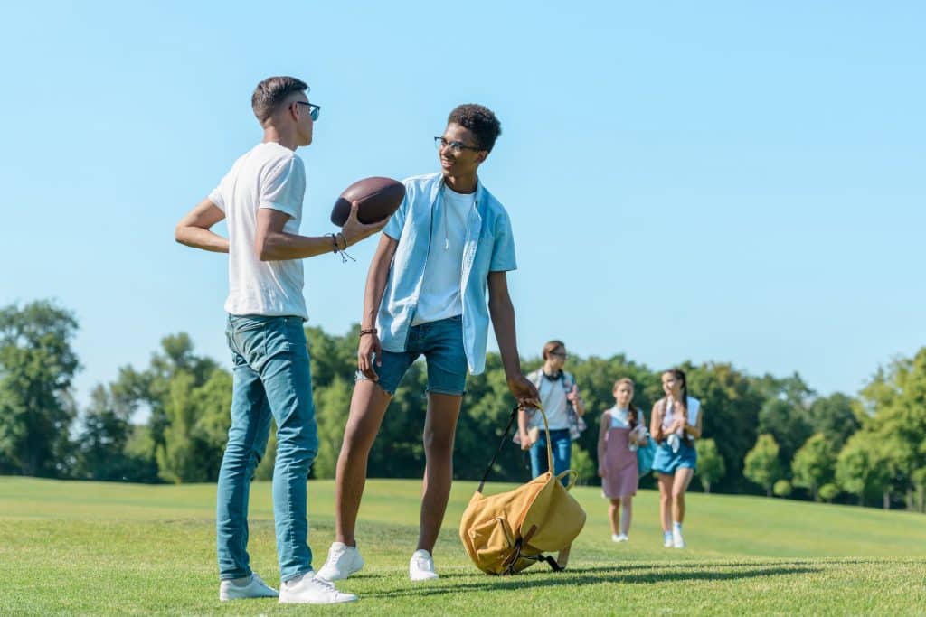 multiethnic boys playing with rugby ball while classmates walking behind in park